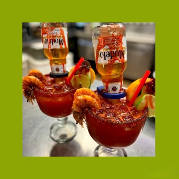 Two Modelo micheladas with shrimp, lime, and a red straw, on a metallic surface.